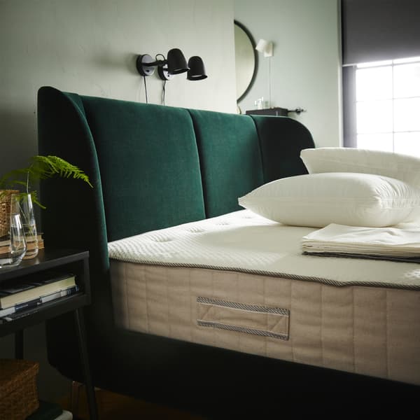 An upholstered bed in green velvet, a white/beige mattress, two white pillows, folded bed linen and two black wall lamps.