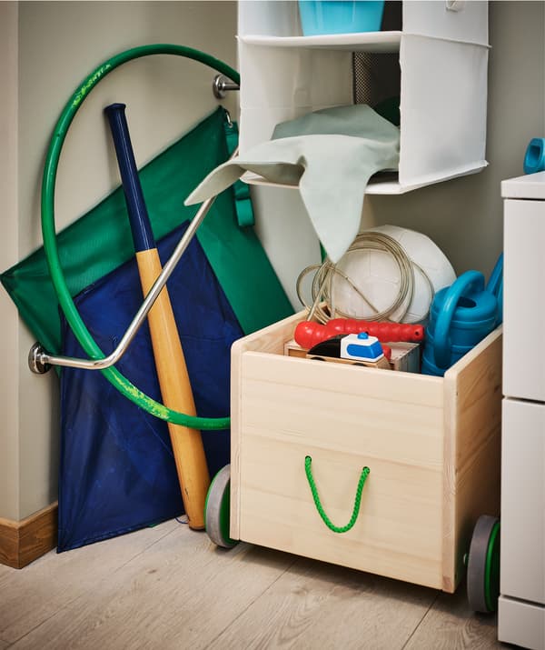 Angolo dell'ingresso con un portasciugamani in diagonale che sostiene degli aquiloni, una mazza e un hula hoop; di fianco, un contenitore giocattoli FLISAT pieno - IKEA