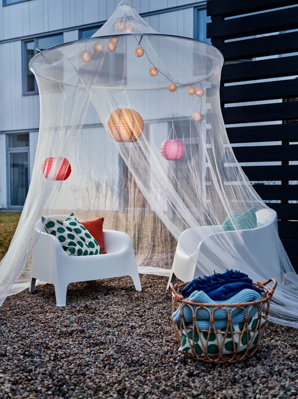 Angolo di un cortile con una rete SOLIG decorata sopra delle poltrone in plastica. Accanto, vari plaid in un cestino SNIDAD.