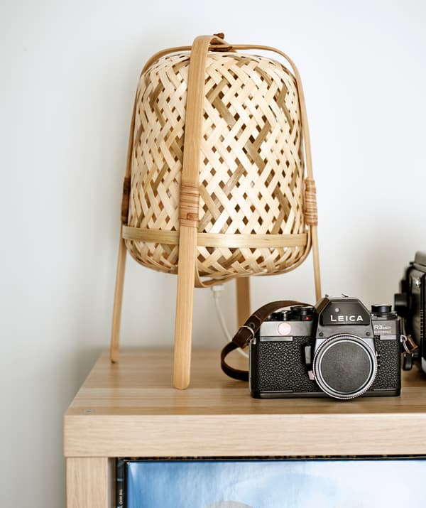 Angolo di un mobile in legno chiaro su cui sono appoggiate una fotocamera vintage e una lampada da tavolo in bambù intrecciato.