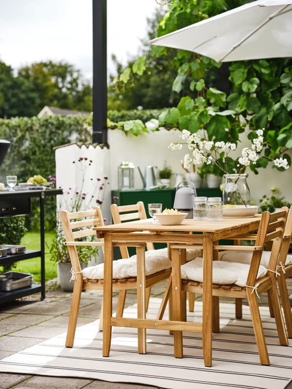 Angolo di un patio circondato dal verde con un tavolo ASKHOLMEN e quattro sedie con braccioli ASKHOLMEN su un tappeto.
