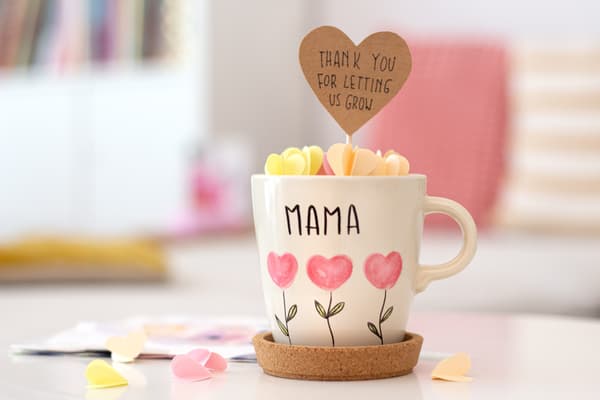 Ansicht der liebevolle handbemalten Tasse für den Muttertag mit einem Gruß aus Papierblumen zum Rausziehen. Die Tasse steht auf einem weißen Tisch 