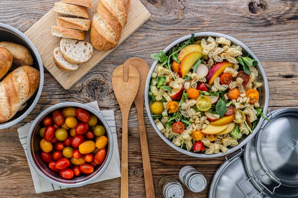 Ansicht von oben auf eine Schüssel mit Nudelsalat, daneben befinden sich Tomaten und verschiedene Brotsorten als Beilage.