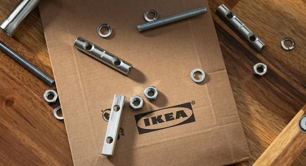 Ant medinio paviršiaus guli kartoninė dėžė su IKEA logotipu ir metalinėmis tvirtinimo detalėmis.