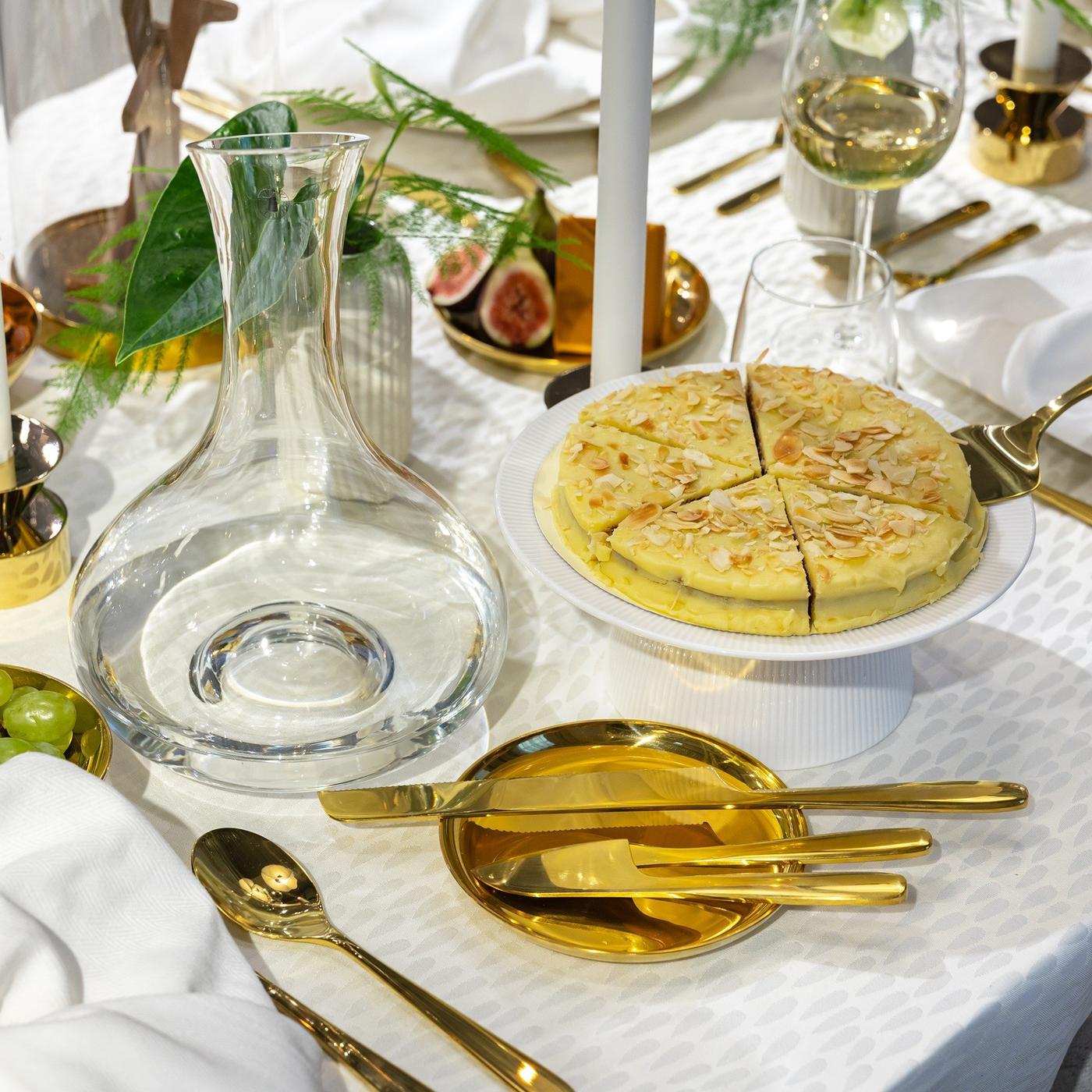 Ant šviesios staltiesės, kartu su auksinės spalvos stalo įrankiais ir šventiniais patiekalais, stovi stiklinis grafinas.