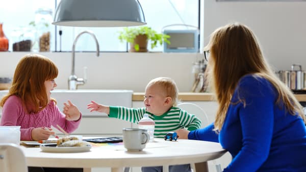 ANTILOP-syöttötuolissa oleva vauva istuu valoisassa keittiössä saarniviiluisen LISABO-pöydän ääressä aikuisen ja toisen lapsen kanssa.