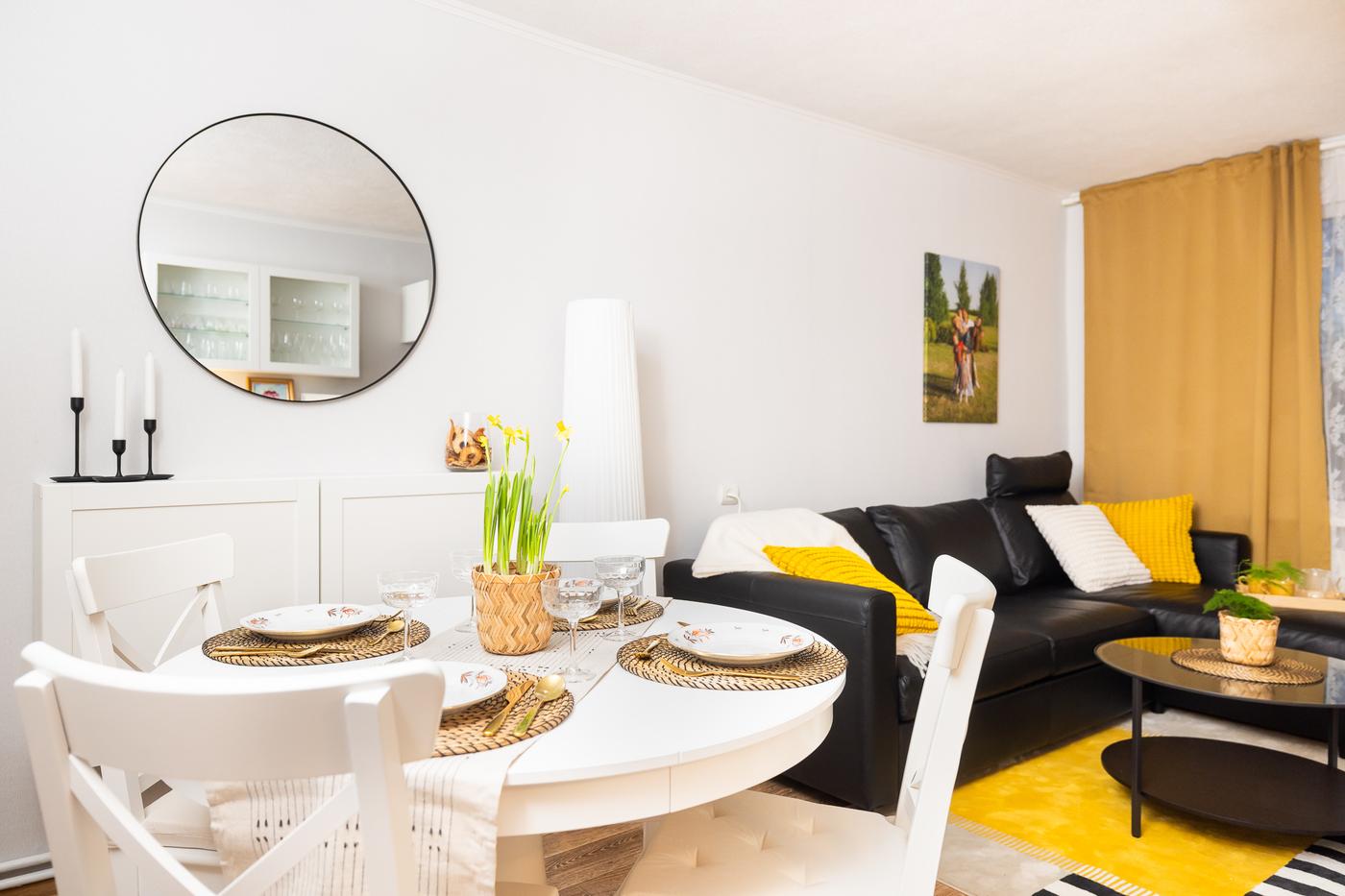 Apaļš ēdamgalds ar baltām krēslu rindām pie sienas ar lielu apaļu spoguli. Telpa izskatās gaiša, sakārtota un vizuāli sadalīta funkcionālās zonās.