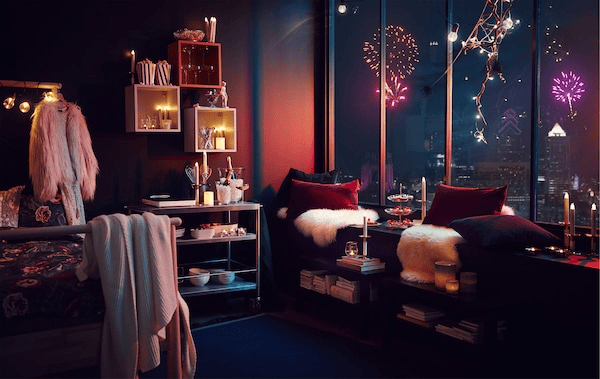 Apartment interior with party-style decorations and lighting; fireworks over a cityscape visible through tall windows.