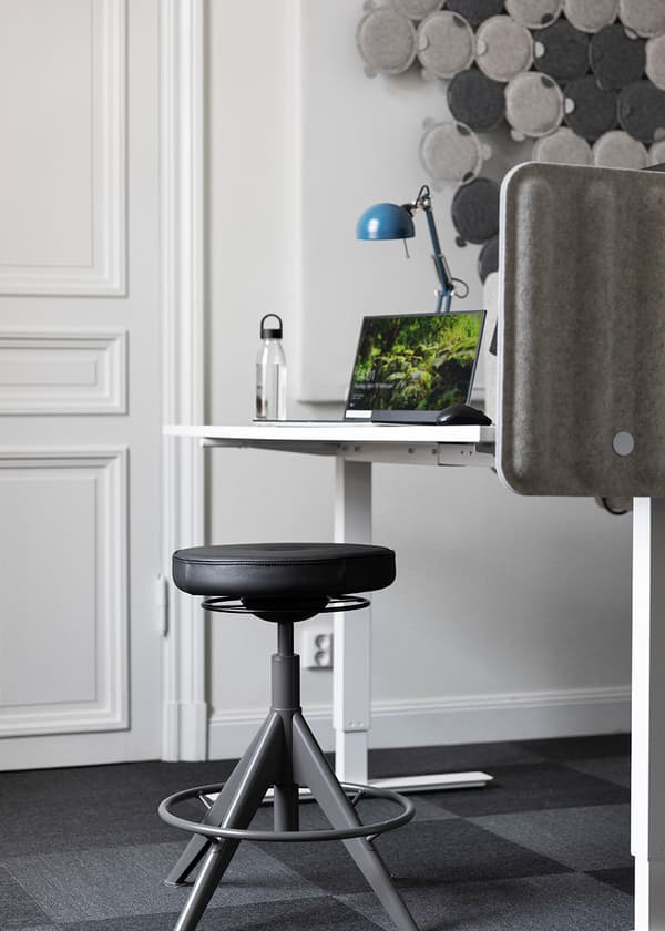 Arbetsplats med svart hög pall, vitt skrivbord, laptop, lampa och avskärmning i ett modernt kontorsutrymme.