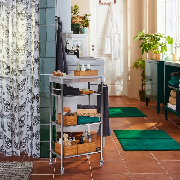 عربة GRUNDTAL ستنلس ستيل تقف بجوار دوش وفوق الرفوف المنتجات اللازمة للاستحمام.