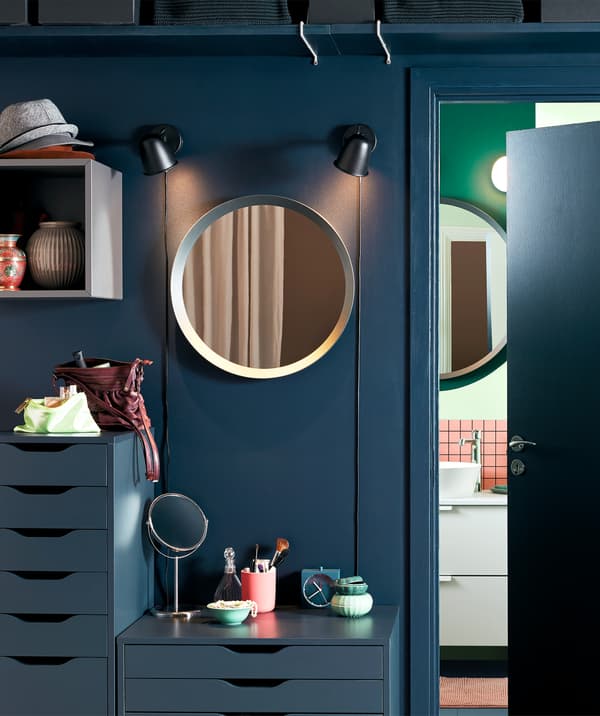 Área exterior de uma casa de banho, decorada como uma versão simplificada de um posto de maquilhagem: espelhos, cómodas e artigos de maquilhagem.