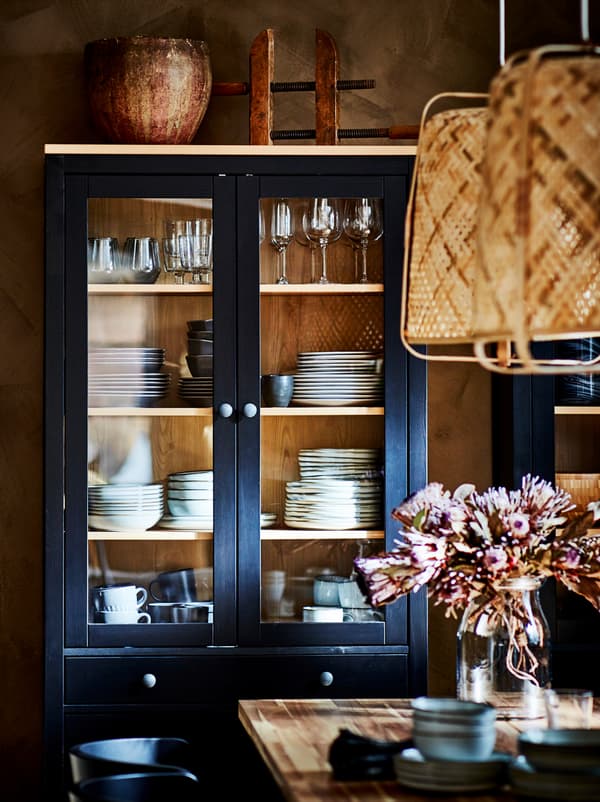 Armário com portas de vidro HEMNES com pilhas de loiça, encostado a uma parede ao lado de uma mesa de refeições.