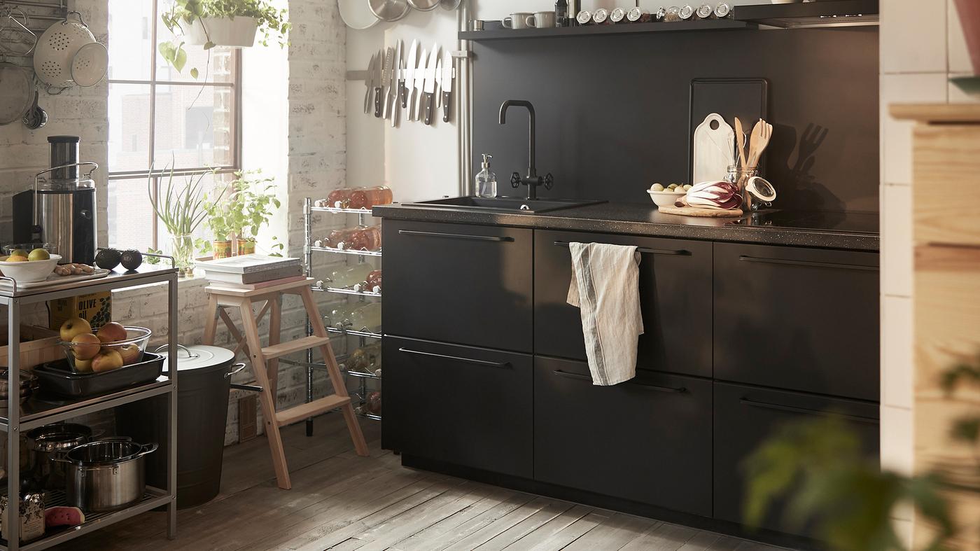 Armários de cozinha em preto com um lava-loiça, um carrinho de metal e utensílios dispostos em calhas junto de uma janela.