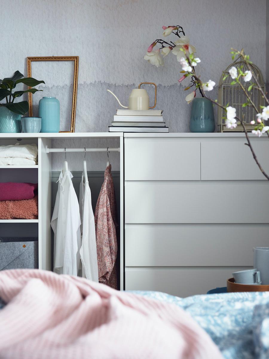 Armoire blanche customisé avec une barre de support intégré sur laquelle trois chemises sont accrochées à l'aide d'un cintre et à côté des rangement avec des habits pliés . Au dessus de cette armoire plusieurs accessoires de décorations