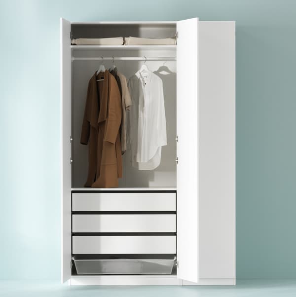 Armoire-penderie PAX blanche contre un mur vert clair, deux portes à charnières ouvertes, où des chemises sont suspendues à une tringle, au-dessus de tiroirs et d’une corbeille en fil.