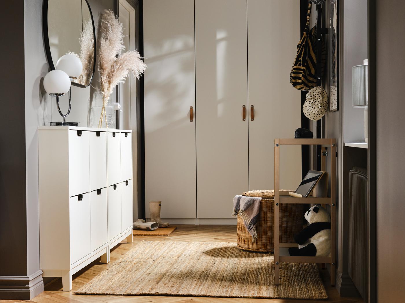 Armoires à chaussures STÄLL sous un grand miroir rond, à côté d’un tapis en jute LOHALS. Armoires-penderies PAX blanches contre le mur adjacent.