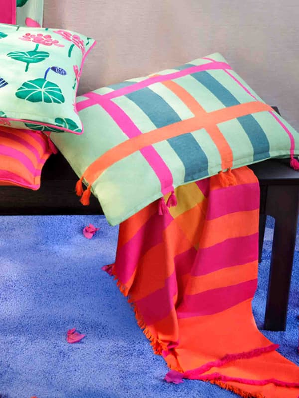 AROMATISK 2024 green cushion cover with flowers, on top of an AROMATISK 2024 throw in bright orange and pink on a bright blue rug.
