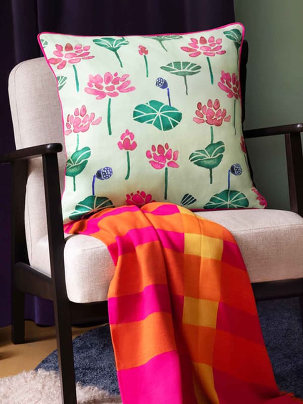 AROMATISK 2024 green cushion cover with flowers, on top of an AROMATISK 2024 throw in bright orange and pink on an armchair.