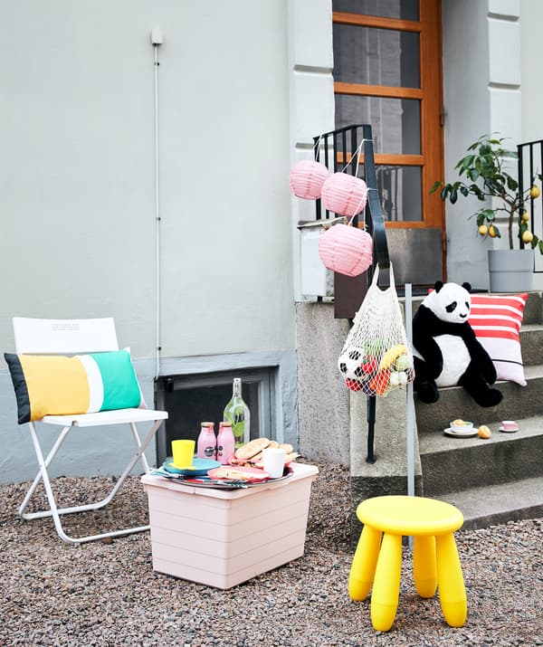 Arrangement for picnic and play outside a building, with seating, a small table, food and drink, toys and decorations.