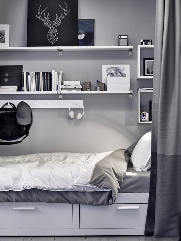 Artwork, books, boxes and more stored on white shelves and hooks above a bed that has drawers beneath.