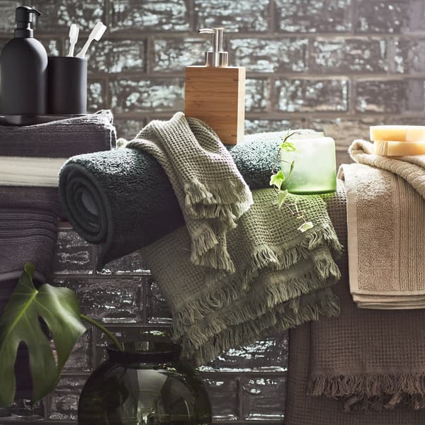 Asciugamani in più colori (beige, grigio chiaro/marrone, grigio scuro e verde) e accessori vari in un bagno con le piastrelle grigie.