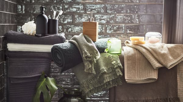 Asciugamani in vari colori (beige, grigio chiaro/marrone, grigio scuro e verde chiaro) e accessori vari in un bagno con le piastrelle grigie.