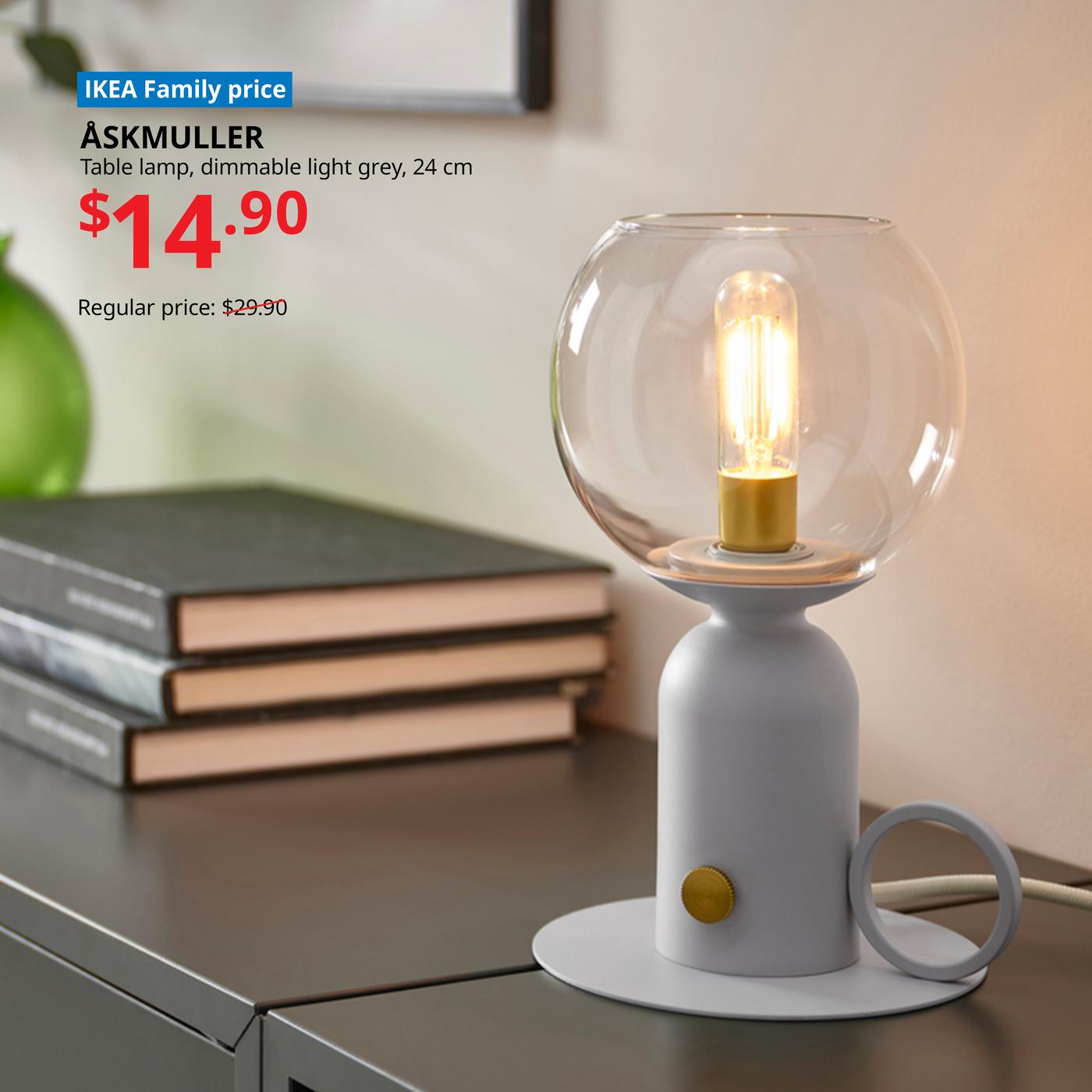 ÅSKMULLER dimmable light grey table lamp with a round glass shade glowing warmly on a bedside table beside stacked books; IKEA Family price $14.90, reduced from $29.90.