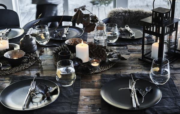 Assiettes noires, couverts en acier, gobelets et centre de table composé d’une bougie bloc allumée et de pommes de pin posées sur un set de table tissé.
