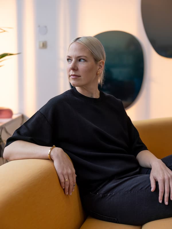 Assise sur un canapé jaune dans son studio, la designer Sabine Marcelis, habillée de noir, tourne sa tête vers la droite.