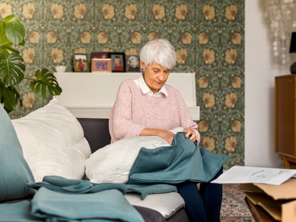 Assise sur un canapé, une femme âgée portant un pull rose clair garnit d’une housse bleue un coussin de canapé.