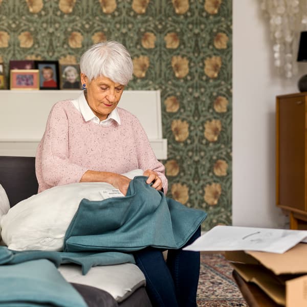 Assise sur un canapé, une femme âgée portant un pull rose clair garnit d’une housse bleue un coussin de canapé.