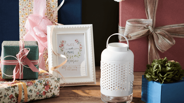 Assorted gift boxes wrapped in colorful paper and ribbons, a framed card with floral design, a white decorative lantern, and a small potted plant displayed on a wooden table.