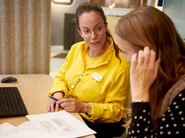 Asztalnál zajló konzultáció számítógéppel, papírokkal és bútortervezettel, sárga munkaruhát viselő személy tollat tart és a tervrajzra mutat.