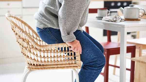 At a white MELLTORP table, a person in jeans and a grey sweater rises from a rattan NILSOVE/NORNA chair with chair pad.