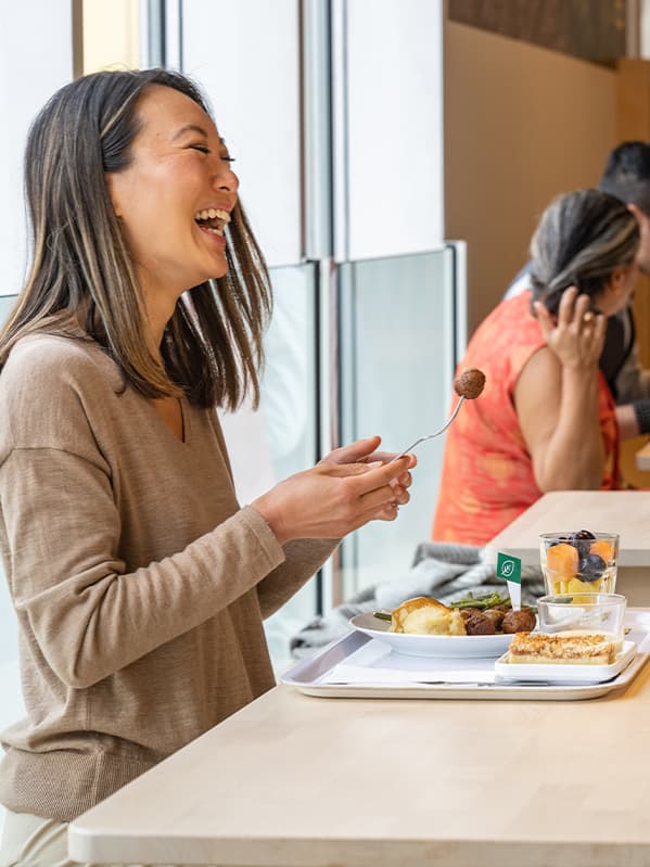 At an IKEA Swedish restaurant, a woman laughs widely while holding a fork with a meatball. In front of her is a tray with a plate of Swedish meatballs and assorted vegetables.