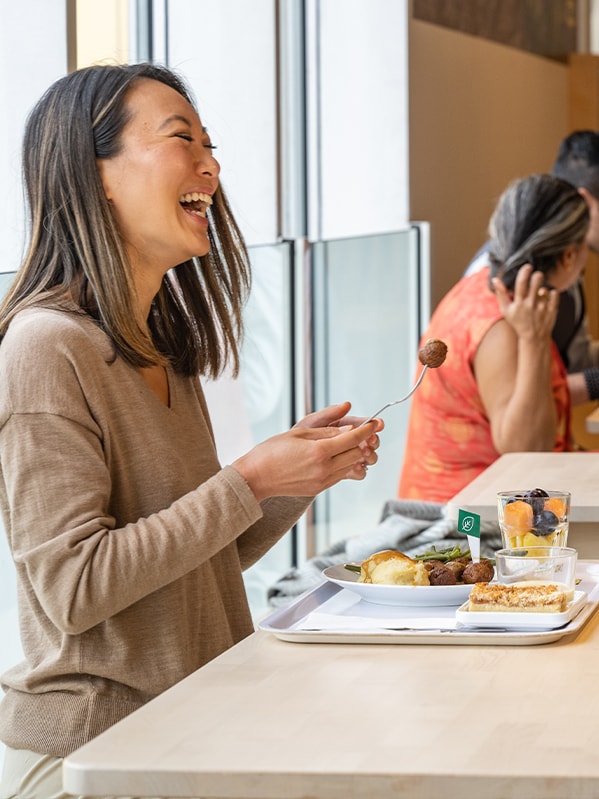 At an IKEA Swedish restaurant, a woman laughs widely while holding a fork with a meatball. In front of her is a tray with a plate of Swedish meatballs and assorted vegetables.