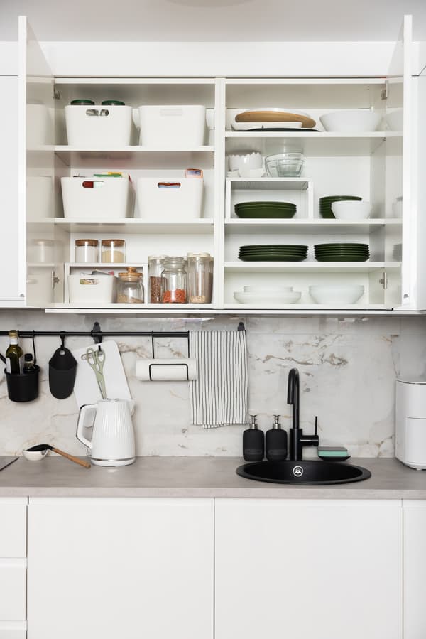 Atvērti augšējie virtuves skapīši ar plauktiem, kuros sakārtoti trauki, kastes un virtuves piederumi.