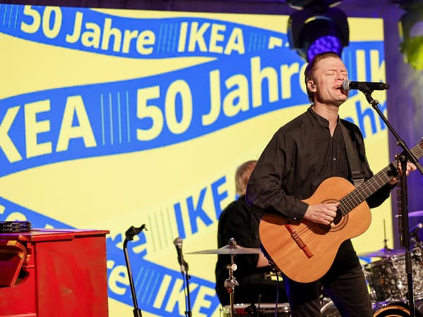 Auf dem Bild sieht man den Sänger der schwedischen Rockband Mando Diao, der Gitarre spielt und singt. Im Hintergrund ist ein großer Bildschirm mit der Aufschrift „50 Jahre IKEA“ in gelben Buchstaben auf blauem Hintergrund zu sehen. Der Sänger trägt ein schwarzes Hemd und wird von einem Schlagzeuger begleitet. Die Szene stammt von der Jubiläumsfeier zum 50-jährigen Bestehen von IKEA in Deutschland.