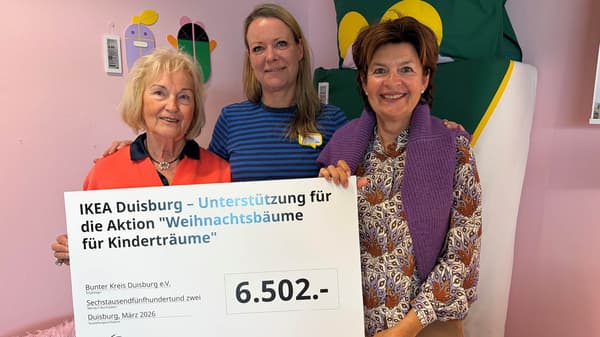 Auf dem Foto stehen drei Personen in einem farbenfrohen Innenraum und halten gemeinsam einen großen Spendenscheck von IKEA Duisburg über 6.502 € für die Aktion „Weihnachtsbäume für Kinderträume“.