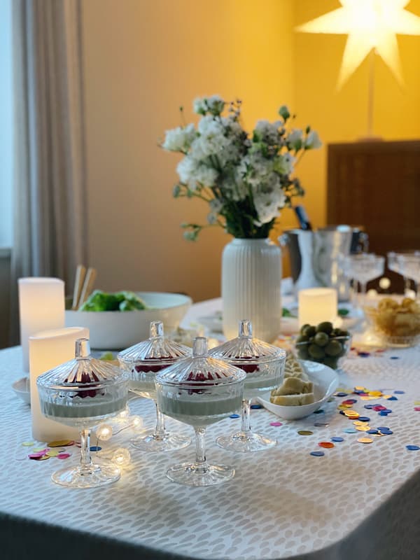 Auf der geschmückten Silvestertafel sind kristallene SÄLLSKAPIG Dessertschalen mit Deckel angerichtet. Darin befindet sich eien weiße Dessertcreme mit dunkelroter Beerensoße. 