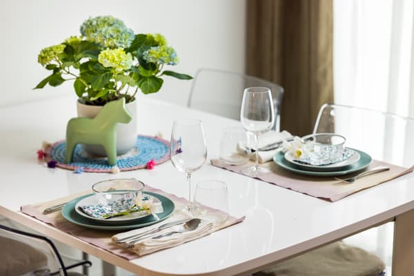 Auf diesem Tisch steht viel buntes Geschirr und jeweils 1 Glas davor. Auf der Seite steht eine weiße Vase mit einer grünen Pflanze.
