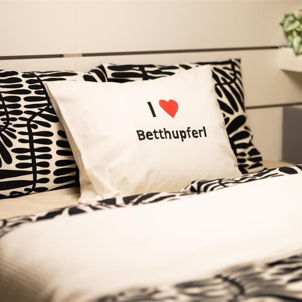 Auf einem Bett liegt ein weißer Polster mit einem Herz. 