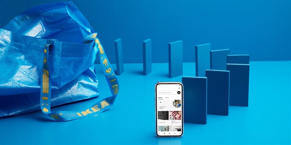 Auf einem blauen Tisch stehen eine FRAKTA Tasche und mehrere Smartphones mit blauen Cover wie Domino Steine aufgereiht und am Ende steht ein Smartphone auf dem man die Startseite der IKEA App sieht.