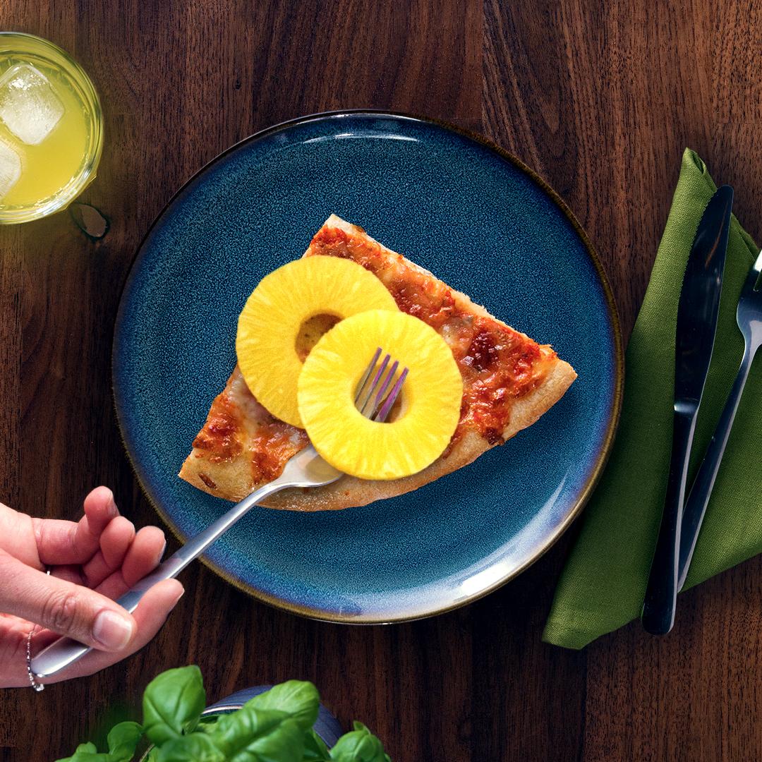Auf einem dunklen Tisch steht ein blauer GLADELIG Teller mit einem Stück Pizza. Darauf liegen zwei Ringe Ananas und jemand fädelt einen Ananasring mit einer Gabel auf.