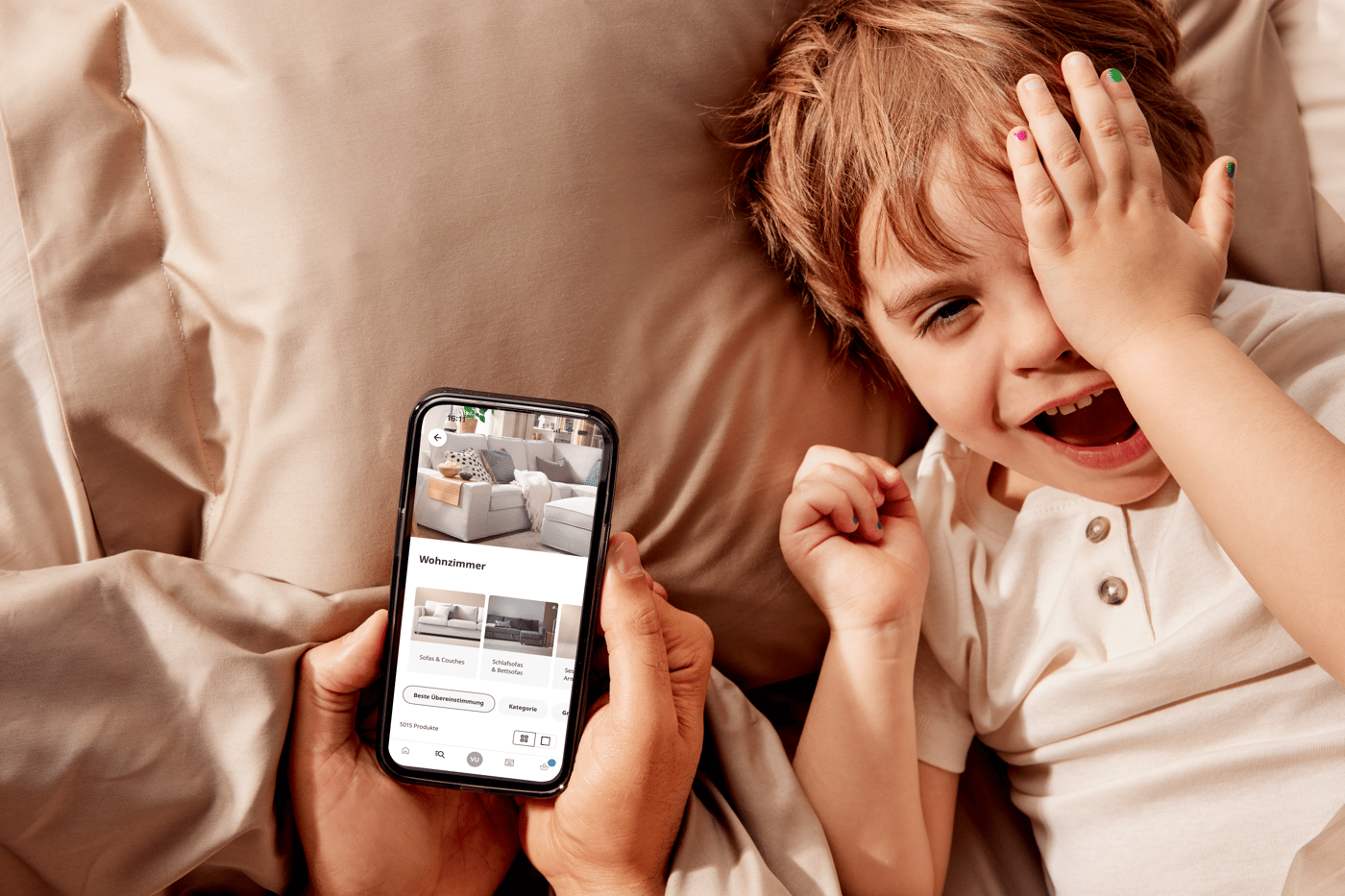 Auf einem Kissen liegt ein lachendes Kind und man sieht wie ein Erwachsener ein Smartphone in den Händen hält und am Bildschirm sieht man die IKEA App