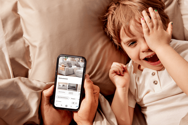 Auf einem Kissen liegt ein lachendes Kind und man sieht wie ein Erwachsener ein Smartphone in den Händen hält und am Bildschirm sieht man die IKEA App