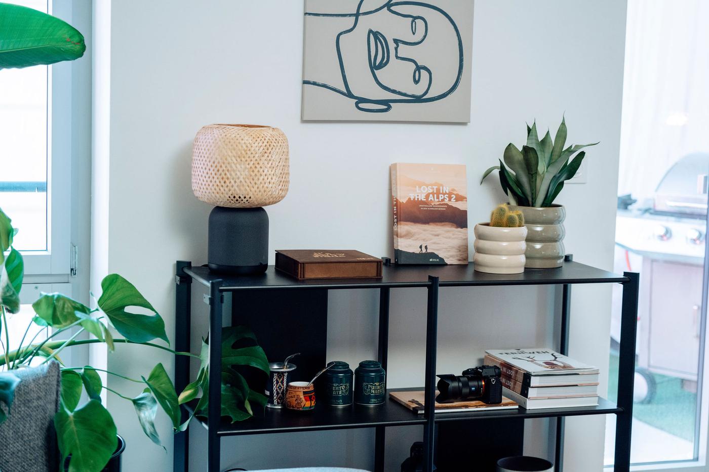 Auf einem offenen Beistell-Regal stehen Pflanzen, eine IKEA SYMFONISK Tischlampe mit Bluetooth-Lautsprecher und mehrere Bücher
