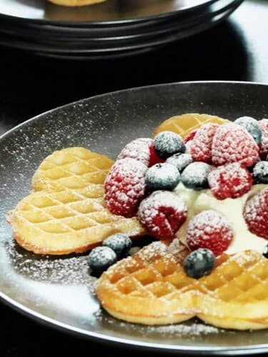 Auf einem schwarzen Teller liegen Waffeln in Herzform und darauf liegen Beeren. 