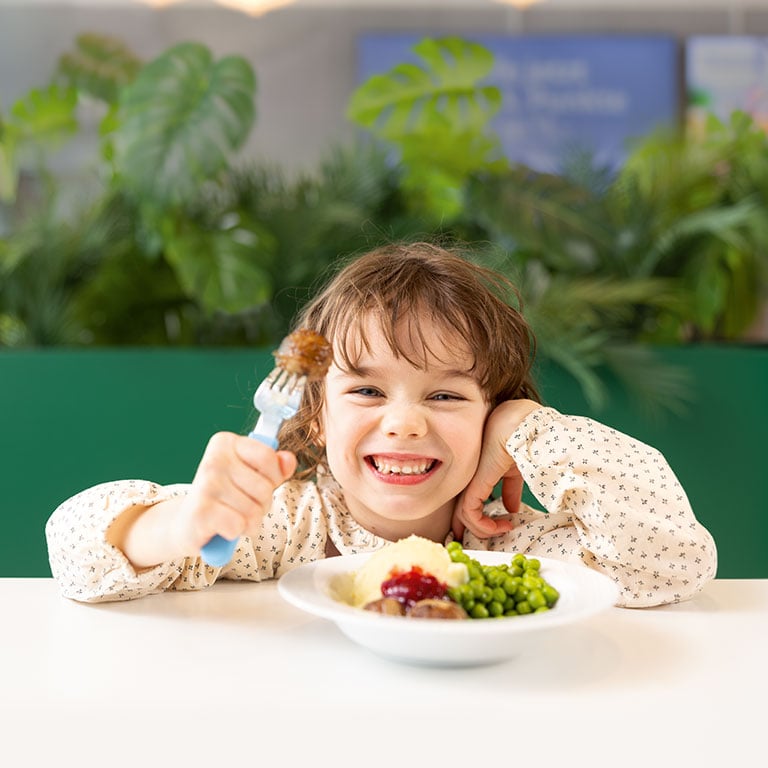 Auf einem Tisch im IKEA Restaurant sitzt ein lächelndes Mädchen vor einem Teller und auf einer Gabel hat sie ein Bällchen aufgespießt und sie lächelt in die Kamera.