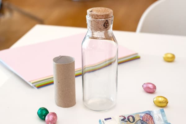 Auf einem Tisch liegen die Materialien: Die Karaffe, eine Klopapierrolle, Bastelpapier, ein paar Schokoeier und ein Geldschein. 